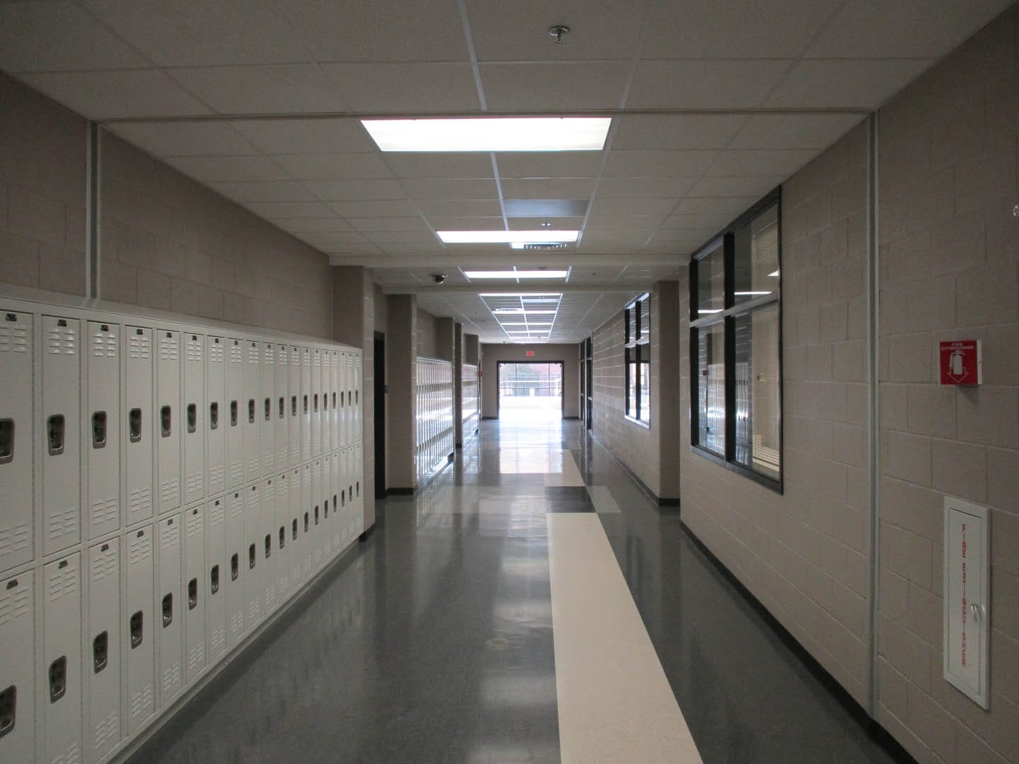 An empty school hallway with beige walls, white lockers on the left, large windows on the right, and a fire extinguisher mounted on the wall.