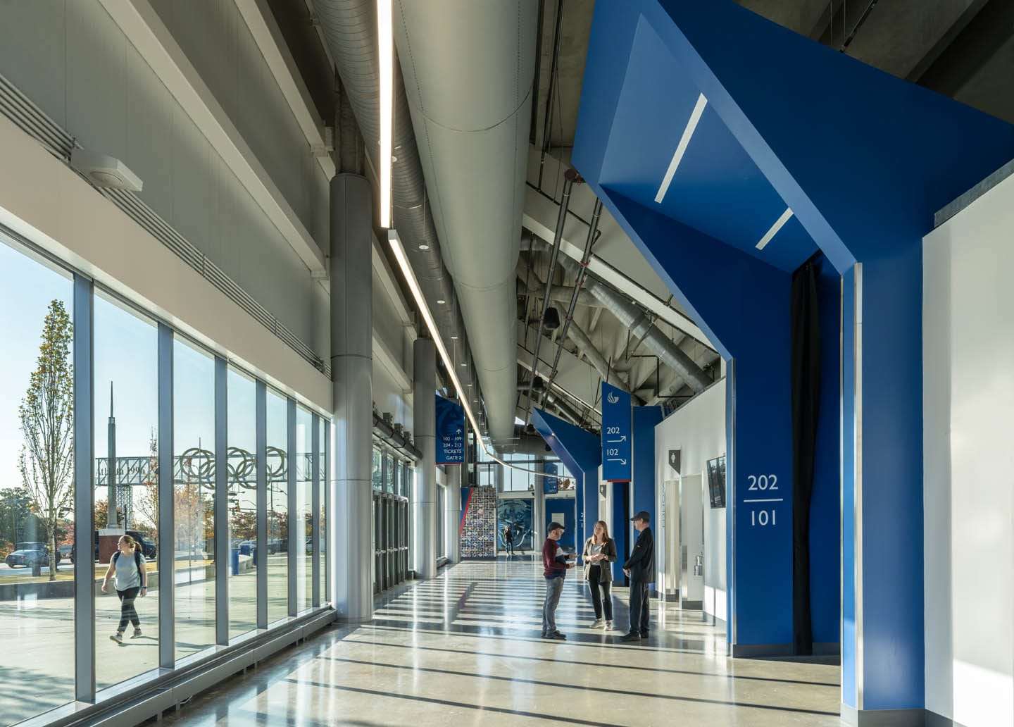 Convocation Center Georgia State University: Interior of a modern building hallway with high ceilings, large windows, and blue structural beams. People are standing and talking near a sign marked "202 101.