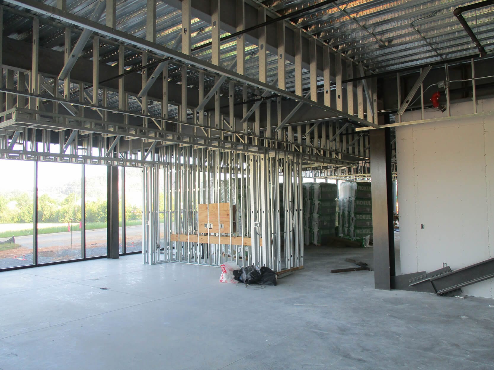 A construction site with metal framing and unfinished walls stands as a blank canvas. Large windows reveal a grassy landscape outside. Building materials are stacked in the background, ready to shape this space into a vibrant centre of innovation and design by Shaw.