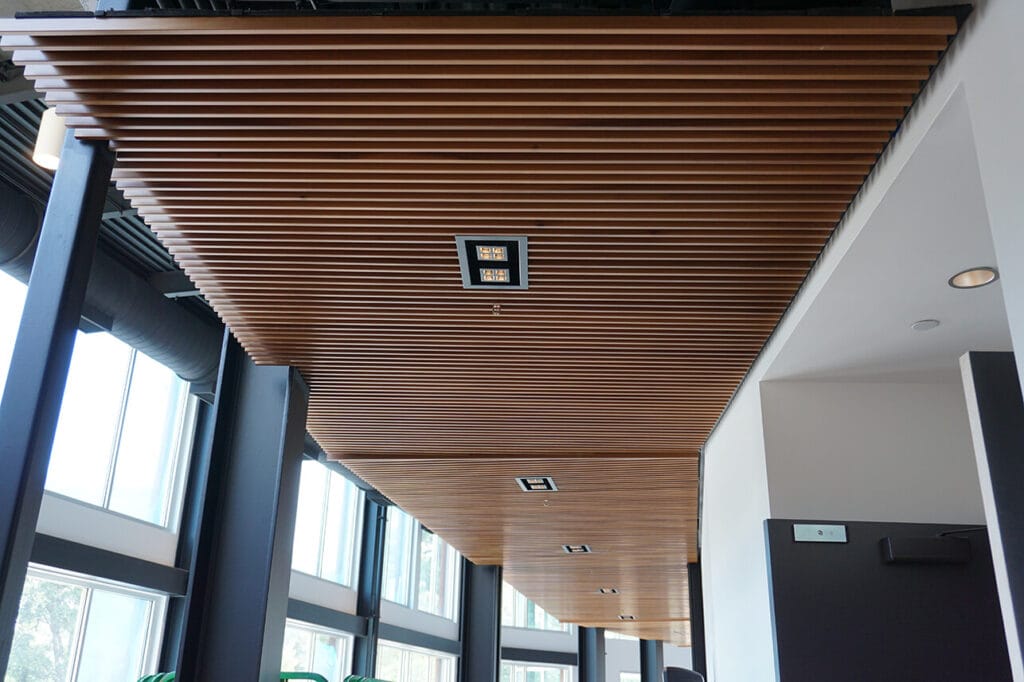 A modern wooden slat ceiling with recessed lighting fixtures in a room with large windows offers an elegant touch while also providing sound dampening benefits.
