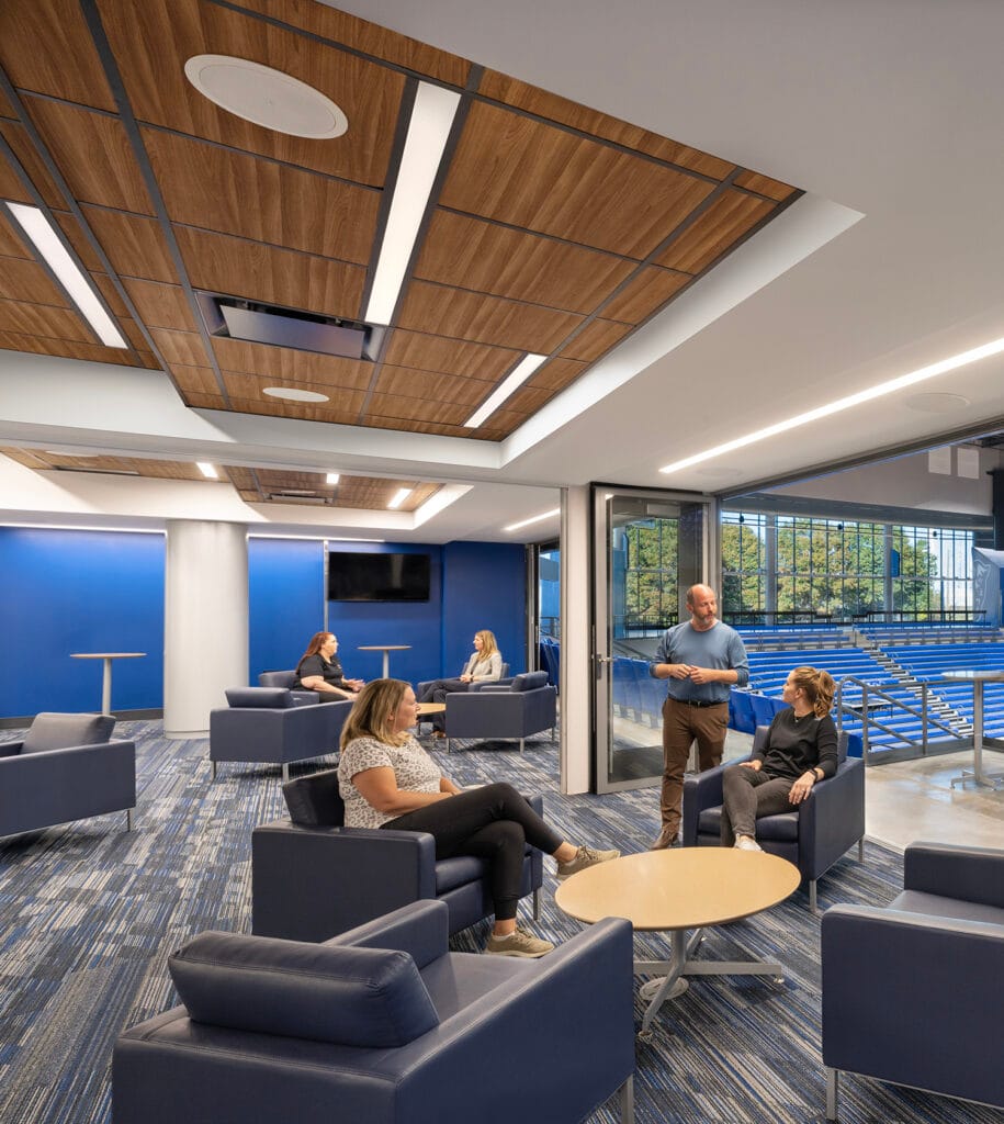 People sitting and conversing in a modern lounge with blue chairs, wooden ceiling panels, and a view of trees outside through large windows, where acoustical ceilings enhance the serene atmosphere.