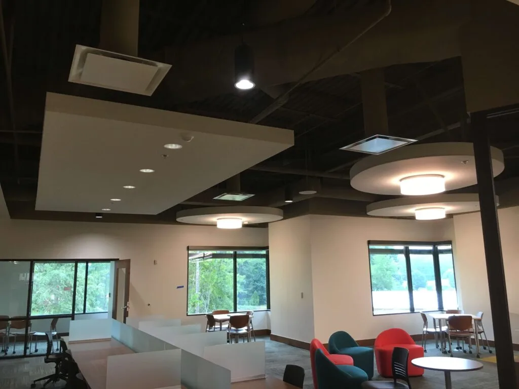 Office space with modern furniture, including desks, chairs, and lounge seating. Large windows provide natural light. The ceiling showcases recessed lighting and exposed ducts, reflecting the sleek style of contemporary commercial construction.