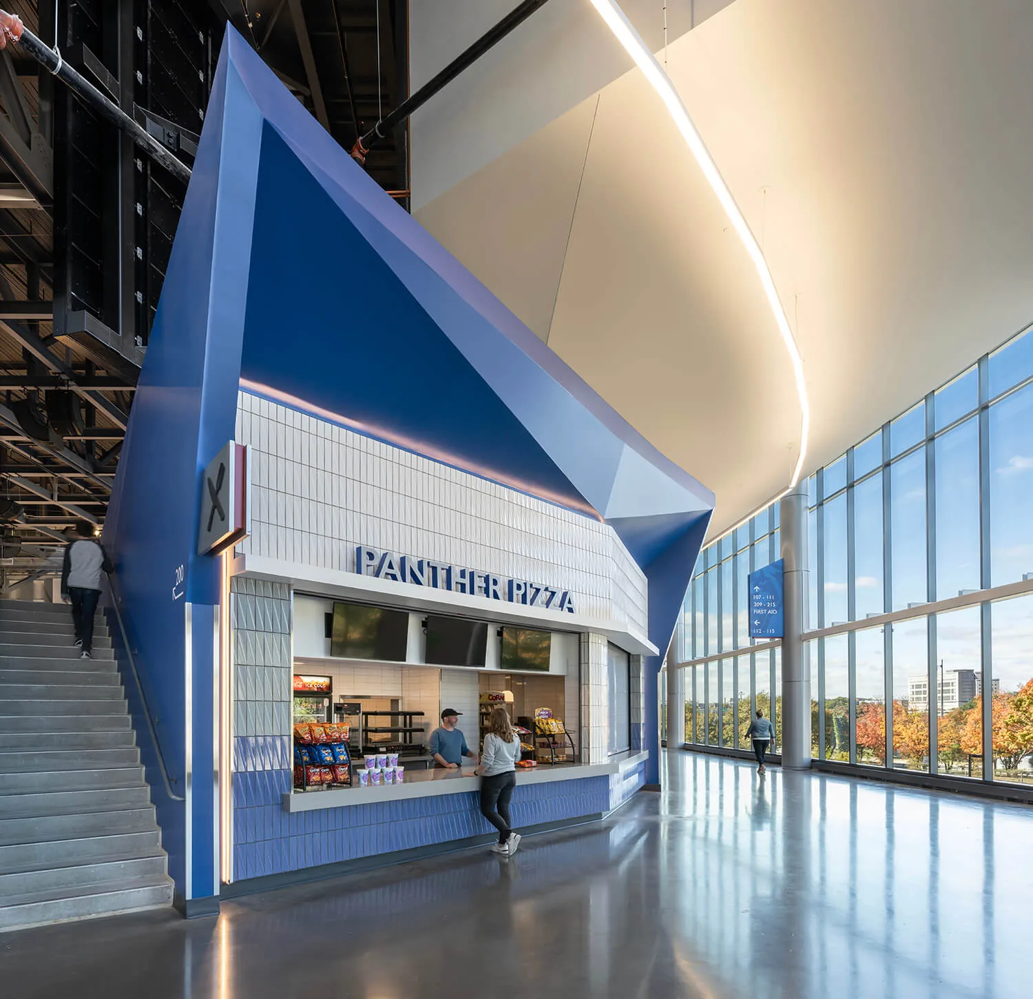 The modern pizza stand, "Panther Pizza," boasts a geometric blue design and is situated beside a staircase and large windows. Nearby, two people engage in conversation, highlighting the area's dynamic commercial construction atmosphere.