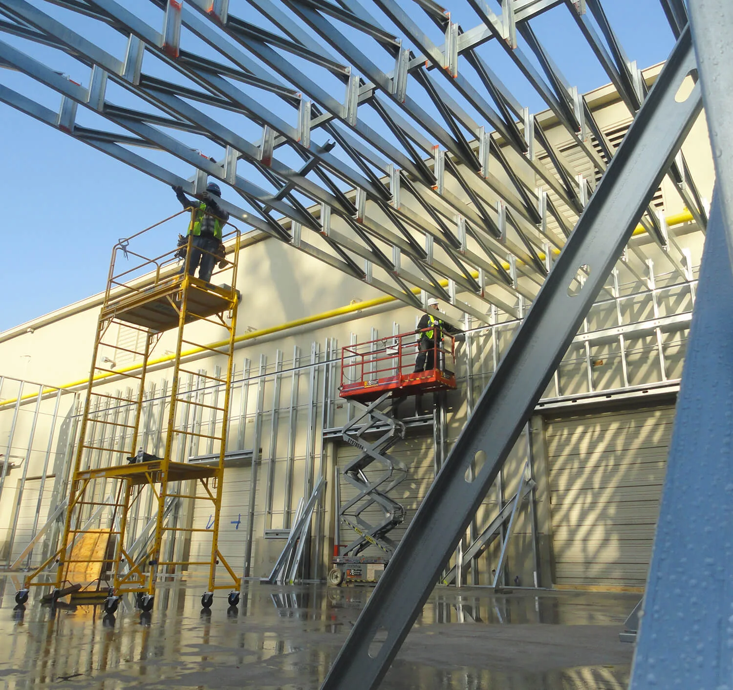 About Us 16 Two construction workers on elevated platforms install a metal frame structure, part of a commercial construction project. The scene is outdoors, with a clear sky reflecting on the wet ground below.