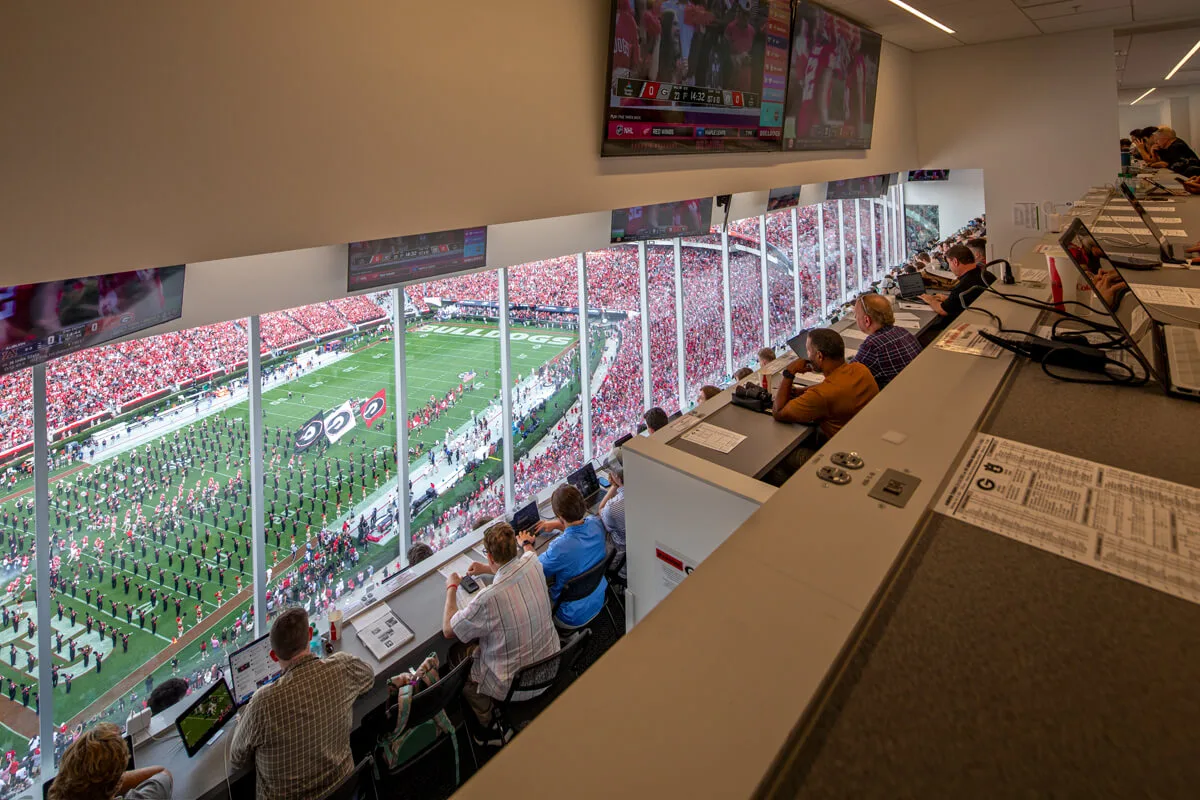 The press box, a marvel of commercial construction, offers an expansive view of the football stadium, where the marching band performs for eager spectators. Inside, reporters diligently work on their laptops, capturing the events unfolding below.