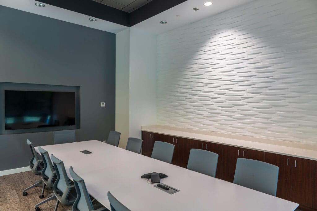 The modern conference room features a white table, gray chairs, a wall-mounted TV, and textured wall paneling—all enhanced by specialty systems. The room is empty and well-lit, ready for dynamic presentations.