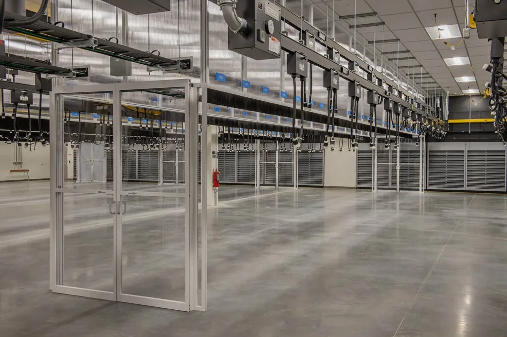 Data Center Work 2 An empty industrial facility with polished concrete floors and overhead equipment serves as a potential data center, featuring a glass double door entrance. Ceiling lights illuminate the spacious, clean interior, providing an ideal work environment.
