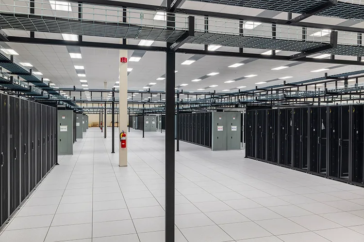 Data Center Work 3 A large, brightly lit data center interior with rows of server racks showcasing the vast amounts of data at work. Overhead cable management ensures organization in this bustling hub. The room has white flooring and a fire extinguisher mounted on a column for safety.