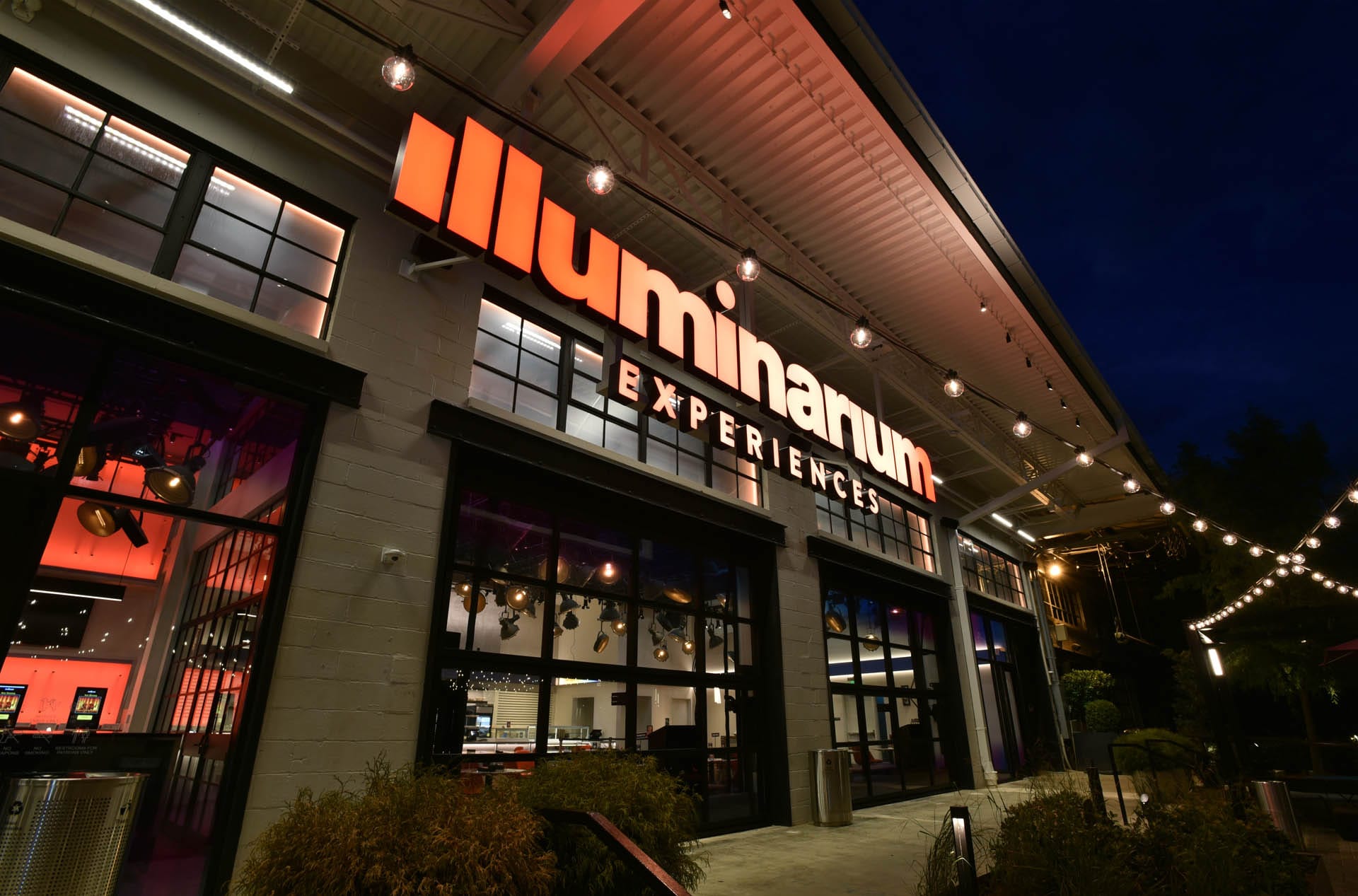 The exterior of the Illuminarium Experiences building at night is captivating, with its illuminated sign and string lights casting a magical glow.