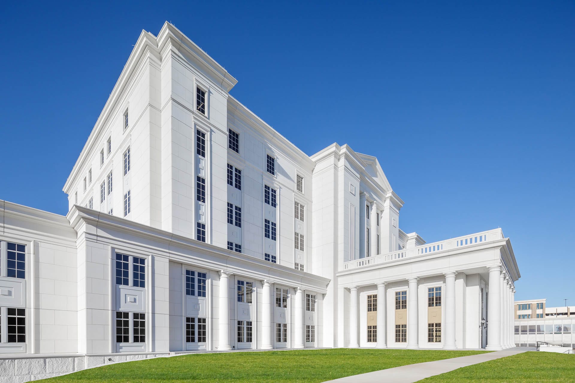 The SCJC Courthouse, a large white neoclassical building with columns and a triangular pediment, stands majestically against the clear blue sky. In the foreground, the plaza's smooth sidewalk winds gracefully through lush green grass.