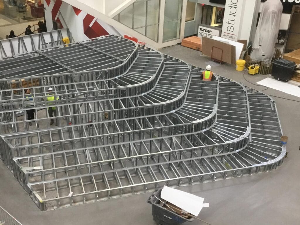 A large, multi-tiered metal framing structure under construction inside a building is surrounded by construction materials and workers in hard hats.