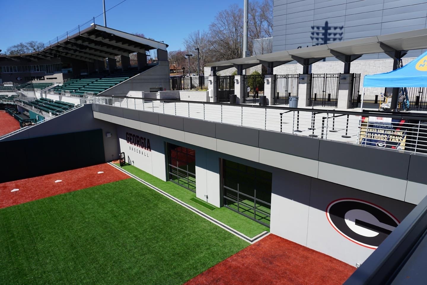 About Us 6 Exterior view of a baseball stadium with Georgia Bulldogs branding, showing green artificial turf, seating areas, and a covered walkway.
