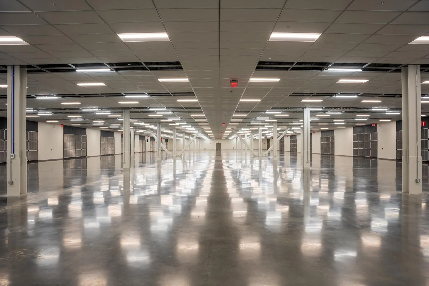 About Us 4 A large, empty industrial warehouse with polished floors, white pillars, and overhead fluorescent lighting stretching into the distance sets the stage for our story—learn more in our About Us section.
