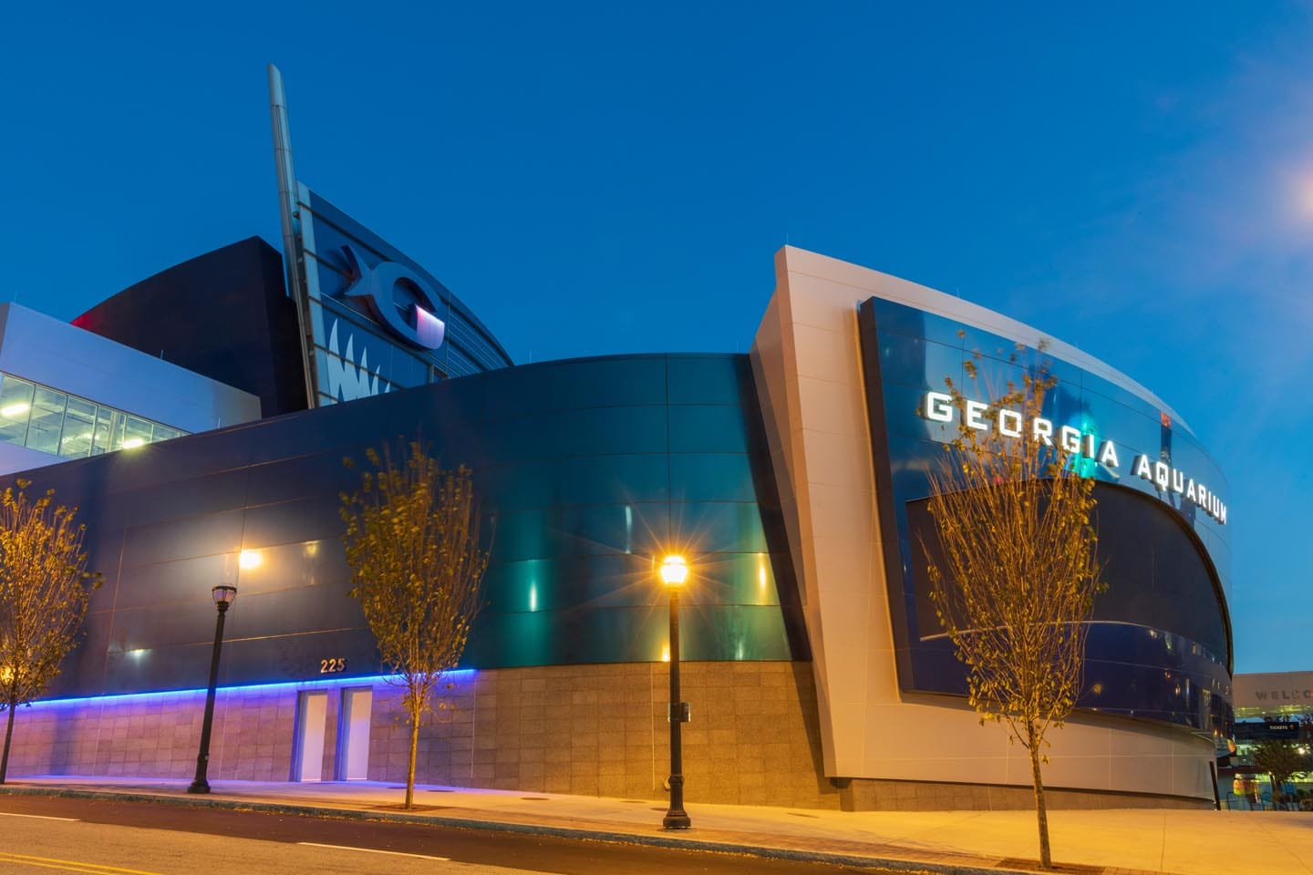 About Us 2 The exterior of the Georgia Aquarium at dusk, featuring modern architecture, illuminated signage that hints at our story, and streetlights along the sidewalk—inviting you to learn more about us.