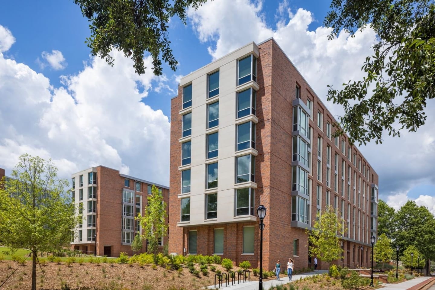 About Us 9 A modern, mid-rise brick apartment building with large windows, surrounded by trees and landscaping, under a partly cloudy sky. Two people walk on a path nearby.