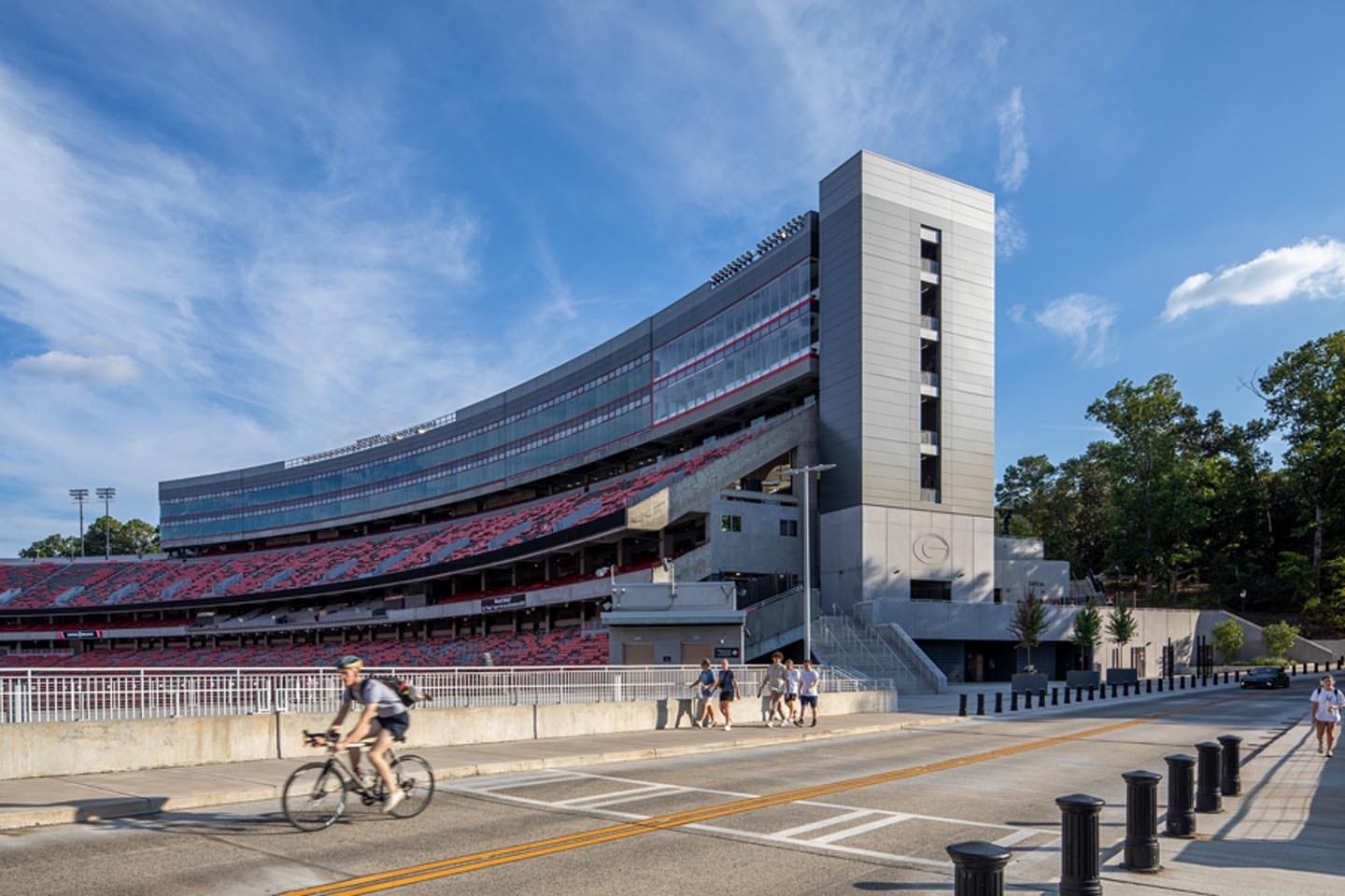 About Us 8 A modern sports stadium with a large, curved grandstand and a tall gray tower, viewed from across a street with pedestrians and a cyclist.
