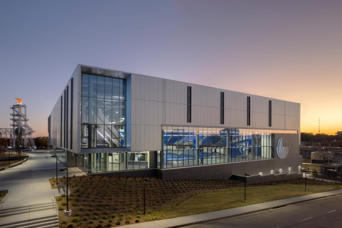 About Us 7 A modern, glass-fronted building with a logo, likely a sports or aquatic center, is shown at dusk with interior lights illuminating a pool area.