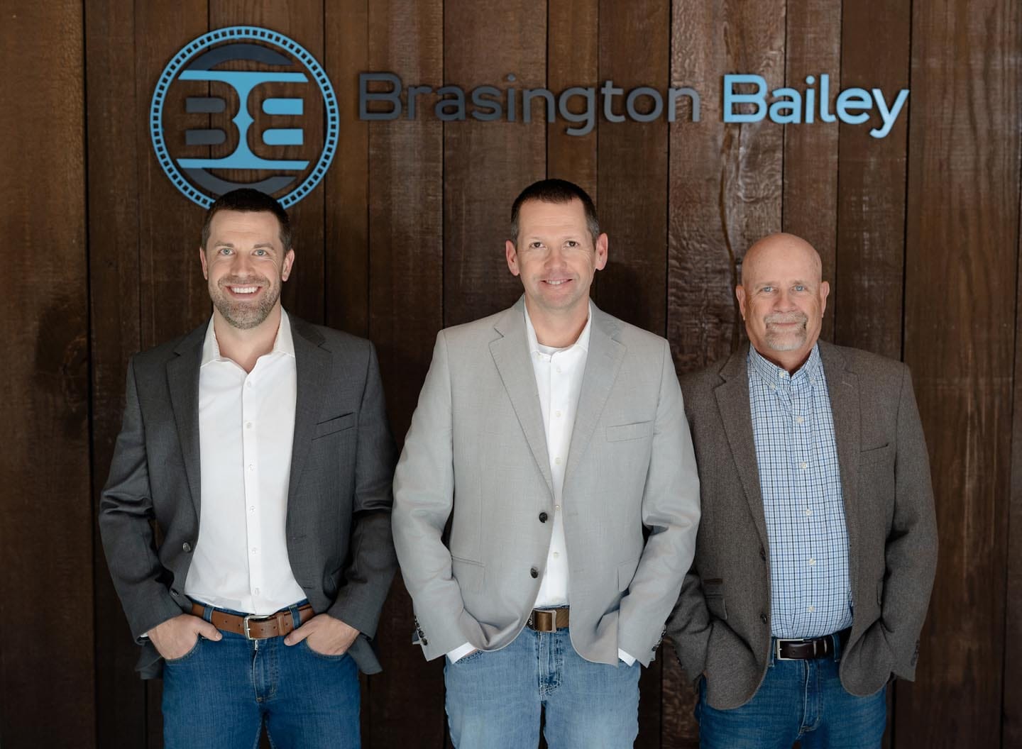About Us 10 Three men in business attire stand in front of a wooden wall with the Brasington Bailey logo and sign.