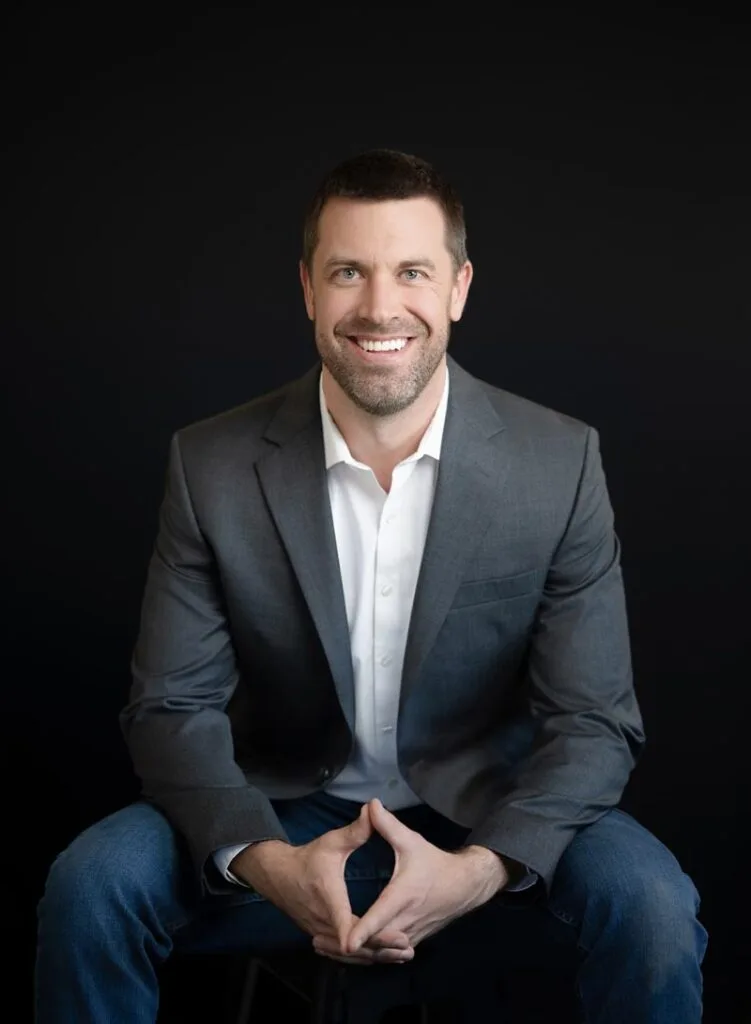 A man with short brown hair, a beard, and a gray suit jacket over a white shirt sits and smiles against a plain dark background.