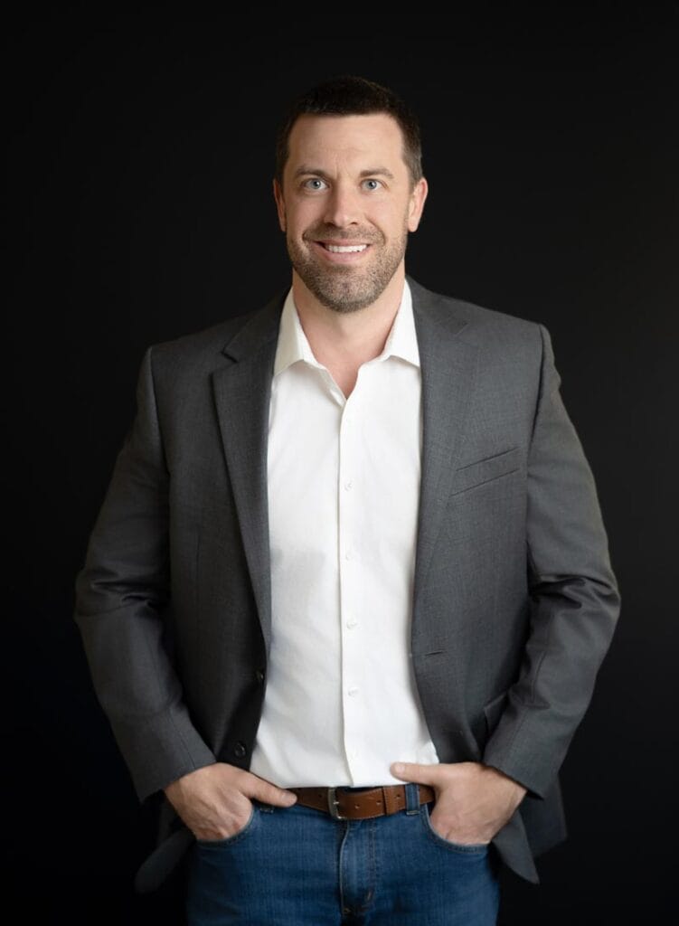 About Us 19 A man in a gray blazer, white shirt, and blue jeans stands against a plain black background with his hands in his pockets, smiling at the camera.