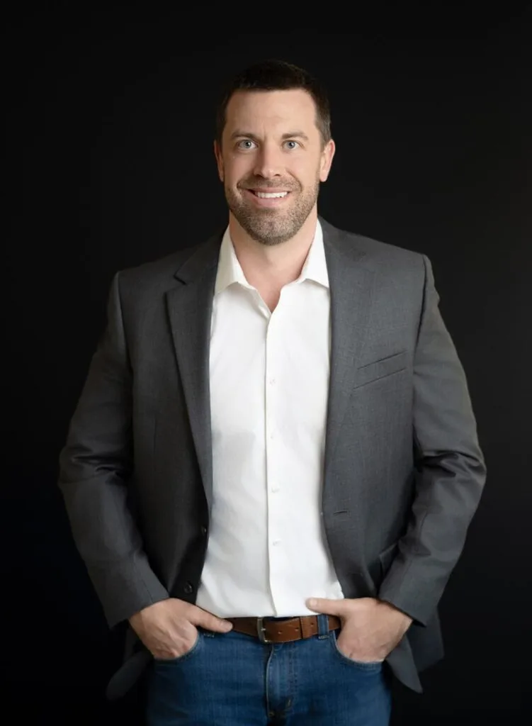 About Us 19 A man in a gray blazer, white shirt, and blue jeans stands against a plain black background with his hands in his pockets, smiling at the camera.