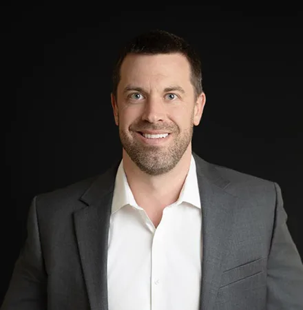 Man with short dark hair and beard, wearing a white shirt and gray suit jacket, smiling in front of a plain black background—perfect for an about us profile photo.