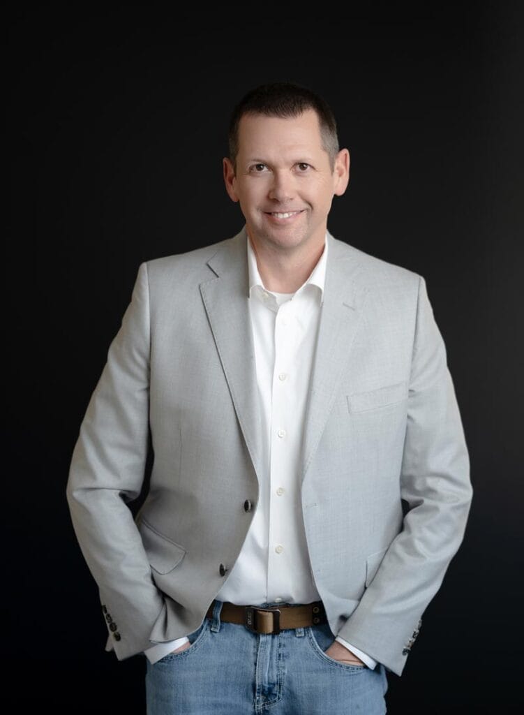 About Us 21 A man in a light gray blazer, white shirt, and jeans stands with hands in pockets against a dark background, smiling at the camera.