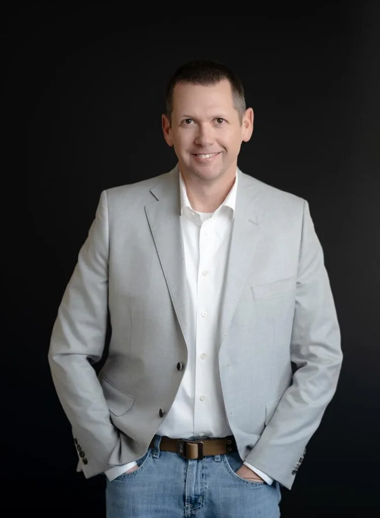 About Us 21 A man in a light gray blazer, white shirt, and jeans stands with hands in pockets against a dark background, smiling at the camera.
