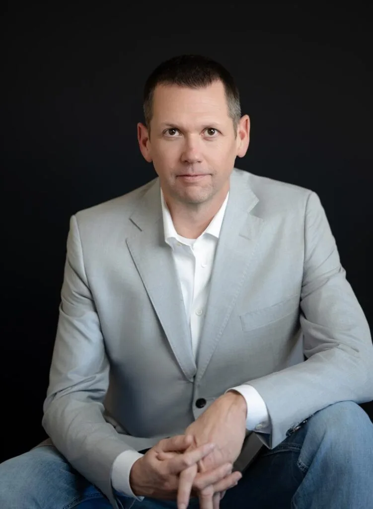 A man in a light gray suit jacket and white shirt sits with his hands clasped, facing the camera against a plain black background.