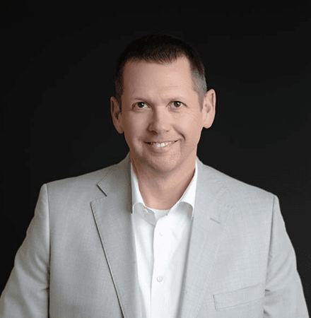 About Us 11 Man wearing a light gray suit jacket and white shirt, smiling, posed against a plain black background—perfect for an "about us" profile photo.