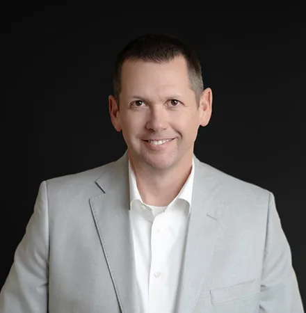 Man wearing a light gray suit jacket and white shirt, smiling, posed against a plain black background—perfect for an "about us" profile photo.