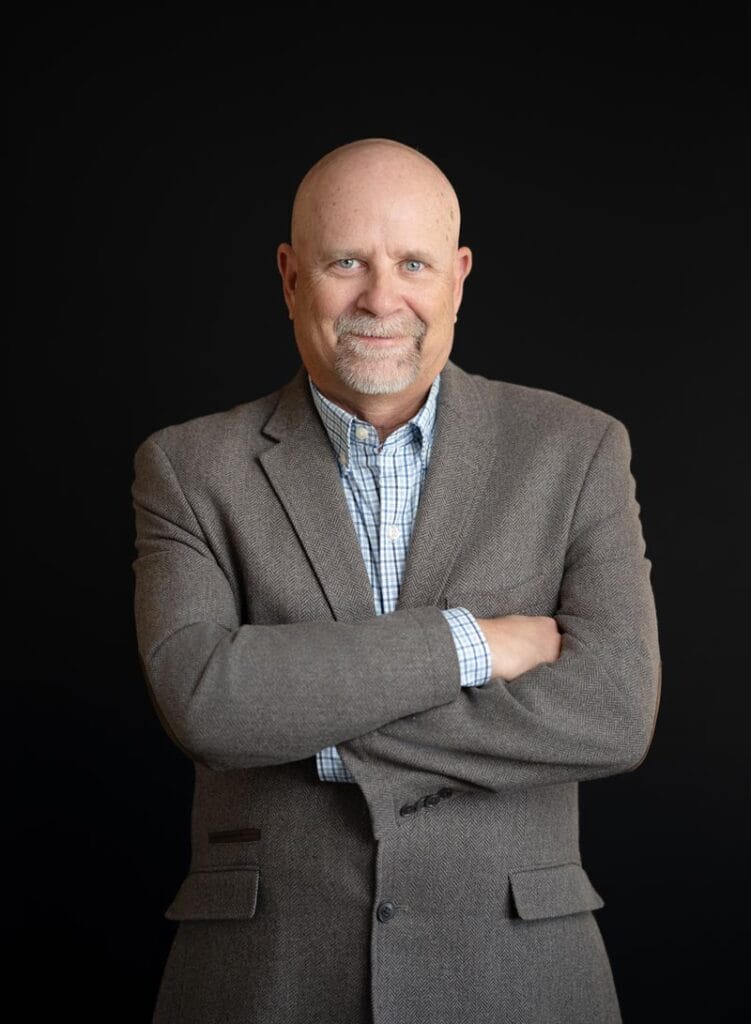 About Us 23 Bald man with a goatee wearing a gray blazer and checked shirt stands with arms crossed against a plain black background.