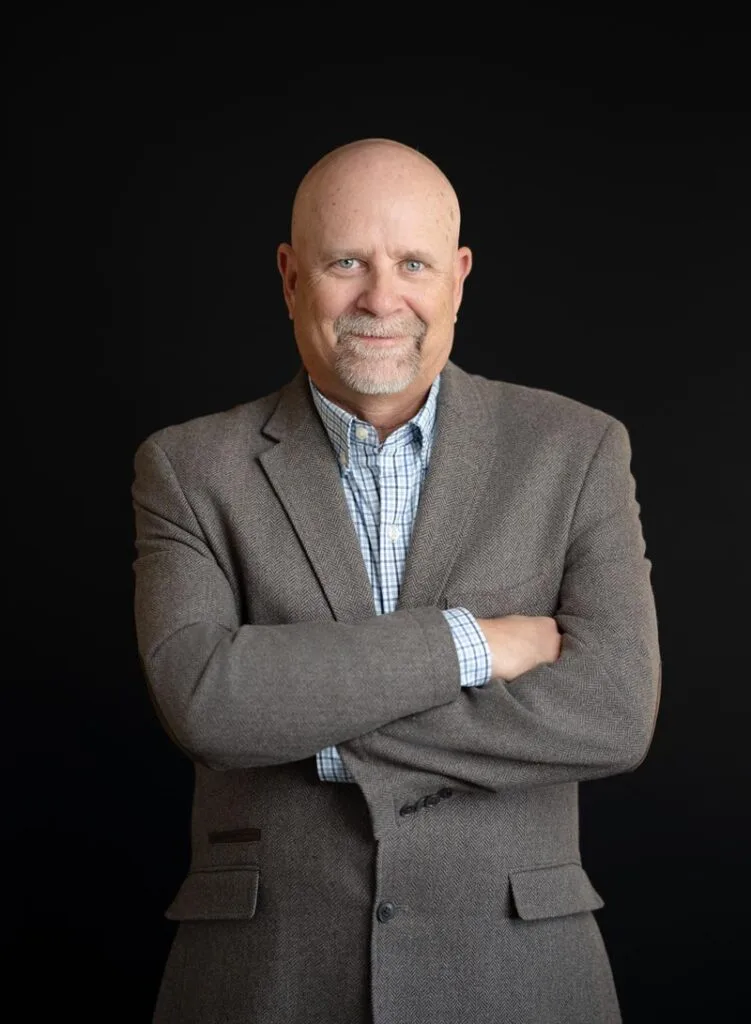 About Us 23 Bald man with a goatee wearing a gray blazer and checked shirt stands with arms crossed against a plain black background.