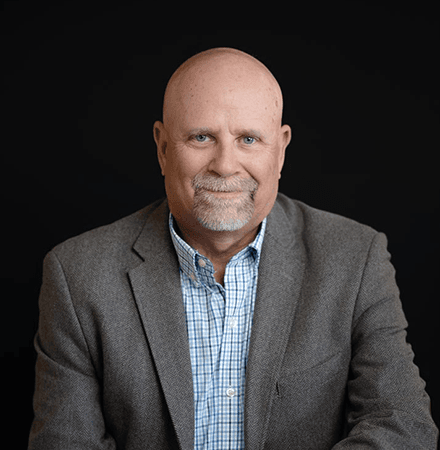About Us 13 Bald man with a gray goatee wearing a gray blazer and checked shirt sits and smiles in front of a plain black background, perfect for an about us page.