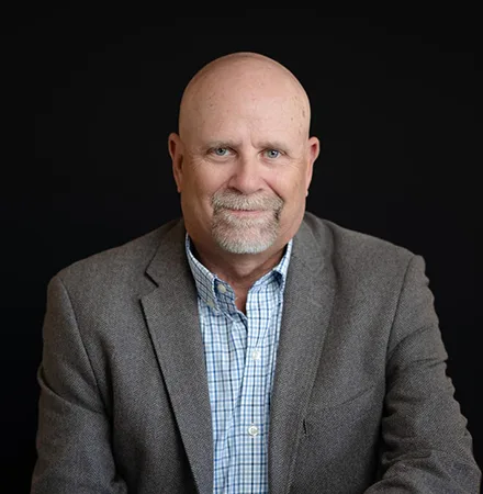 Bald man with a gray goatee wearing a gray blazer and checked shirt sits and smiles in front of a plain black background, perfect for an about us page.
